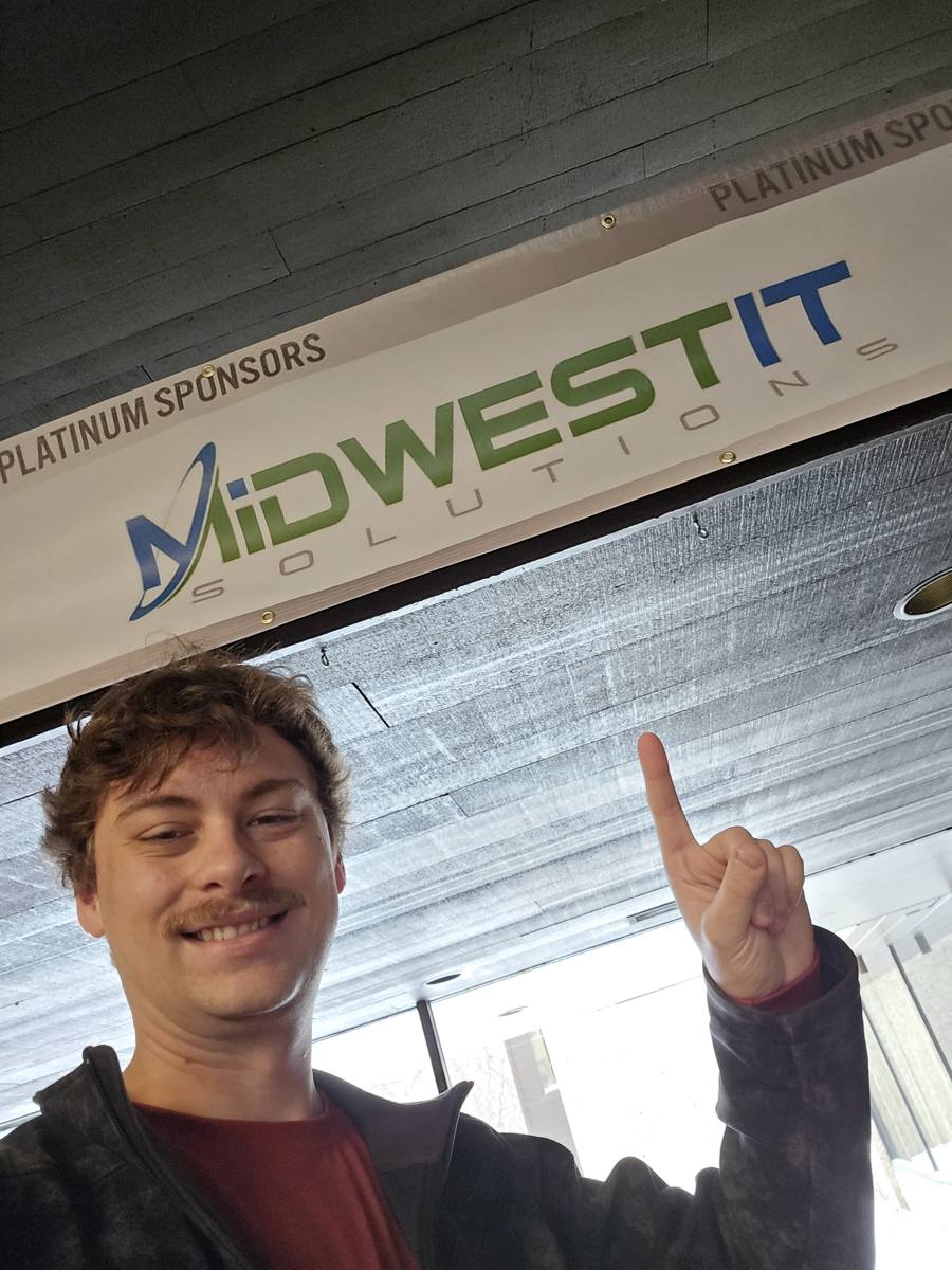 One of the Midwest IT team pointing at the company logo on a platinum sponsors banner